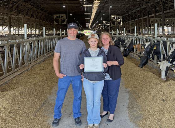 Three people in cow barn, young woman in the middle is holding an award certificate