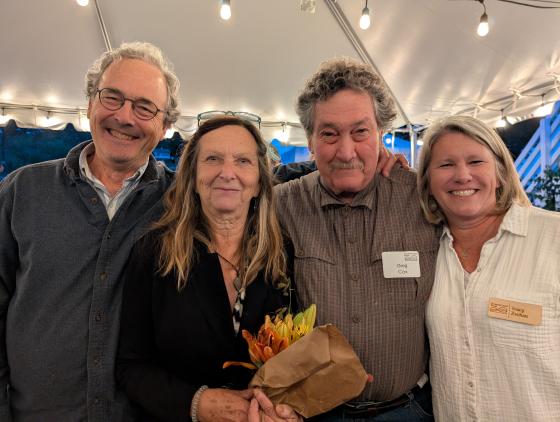 Two women and two men smile at camera, standing under festive lights and white tent (photo of award winner Greg Cox and his wife, Gay Cox)