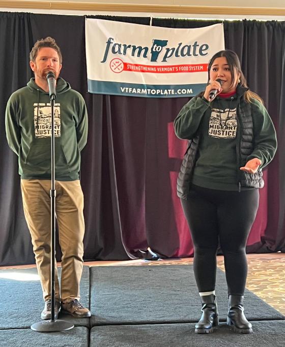 Thelma Gomez of Migrant Justice speaks into a microphone while sharing a personal story with another storyteller on stage at the 2023 Farm to Plate Gathering.